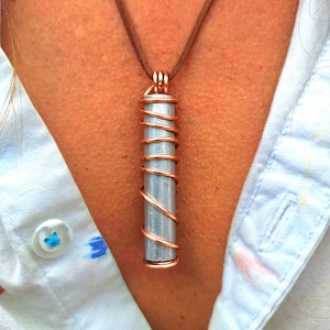 May include: A pendant necklace with a silver-coloured cylindrical stone wrapped in copper wire, suspended from a brown cord. The necklace is worn over a white button-down shirt. The pendant is approximately 5 cm long.