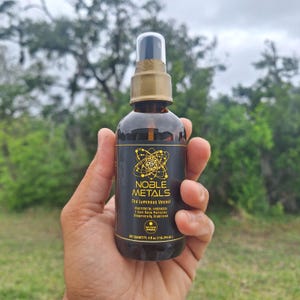 May include: A brown glass spray bottle held in a hand, with a gold cap and a black label. The label reads "NOBLE METALS The Luminous Vessel" with additional text. The bottle contains 4 fl oz (118.294 ml).
