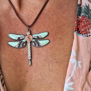 May include: A dragonfly pendant necklace with a brown suede cord. The dragonfly has light blue wings with black and silver detailing. The body is silver with clear crystals and a copper-colored spiral center.