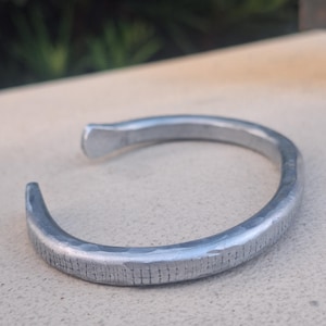 May include: A silver-coloured, open cuff bracelet. The metal bracelet has a textured surface with a hammered appearance. The ends of the bracelet are slightly flattened and curved, creating an open design. The bracelet is a simple, yet stylish accessory.