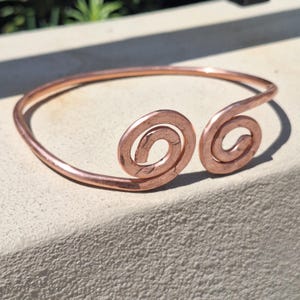May include: A copper cuff bracelet featuring a polished metallic finish and a hammered texture. The design consists of a single continuous band that curves into two symmetrical, circular spiral terminals at the open front of the piece.