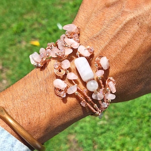May include: A multi-strand wire-wrapped bracelet featuring pale pink tumbled gemstone chips, small clear crystal beads, and a central cylindrical white stone bead. The structure is composed of metallic copper-toned wire coiled into decorative loops.