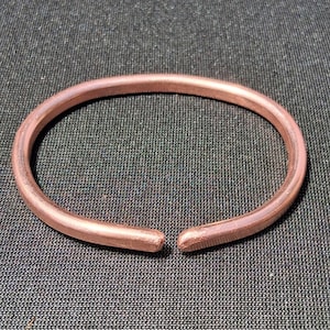 Pode incluir: Uma pulseira circular de cobre com um acabamento metálico liso e uma pequena abertura na parte frontal. A banda tem uma espessura consistente e bordas arredondadas, exibindo o tom castanho-avermelhado característico do metal.