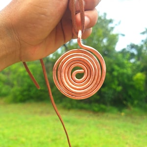 Peut inclure: Un pendentif circulaire fabriqué à partir d'une seule pièce continue de fil de cuivre poli, formé en un motif en spirale serré. Le pendentif est suspendu à un cordon en cuir marron simple, présentant une finition métallique et une texture lisse.