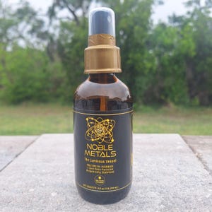 May include: A brown glass spray bottle with a gold cap and black label. The label reads "NOBLE METALS The Luminous Vessel" with additional text. The bottle contains 4 fl oz (118.294 ml) of liquid. The background is blurred green foliage.