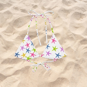 Könnte beinhalten: Ein weißes Bikini-Oberteil mit einem sich wiederholenden Muster aus bunten Seesternen in den Farben Pink, Blau, Gelb und Grün. Das Kleidungsstück verfügt über dreieckige Cups mit verstellbaren Bindebändern am Nacken und Rücken.