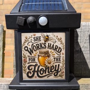 May include: A black solar-powered outdoor light fixture featuring a square decorative panel. The panel displays a honey jar illustration, a bee, a daisy, and the text "SHE WORKS HARD FOR THE HONEY" in black lettering against a cream background.