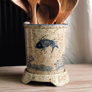 May include: A ceramic utensil holder with a speckled beige finish and a blue fish design. The holder has a blue rim and sits on a matching base. Wooden spoons are inside the holder.