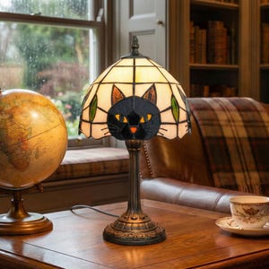 May include: A stained glass table lamp with a black cat design. The lamp features a cream-colored shade with a black cat face, orange nose, and yellow eyes. The base is bronze-colored and ornate. The lamp is on a wooden table.