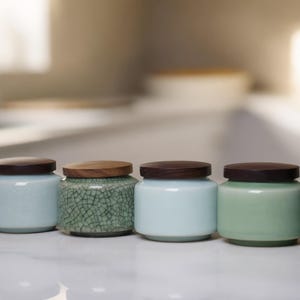Peut inclure: Ensemble de six pots en céramique avec couvercles en bois. Les pots sont de différentes nuances de vert et de bleu, certains avec un glaçage craquelé. Ils sont disposés en ligne sur une surface blanche, design minimaliste.
