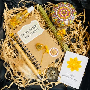 Op de afbeelding: Een geschenkdoos met een notitieboek met de tekst "Every single day counts", een pen, een klein flesje olie, wierook, een kaars en een kaart met de tekst "I radiate personal power".
