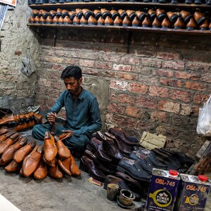 K&ouml;nnte beinhalten: Ein Schuhmacher sitzt auf dem Boden in einer Werkstatt, umgeben von zahlreichen hell- und dunkelbraunen Lederschuhen, Schuhleisten und zwei Metalldosen mit der Aufschrift OSL-745 Power Bond. Regale voller Lederschuhe s&auml;umen die Ziegelwand.
