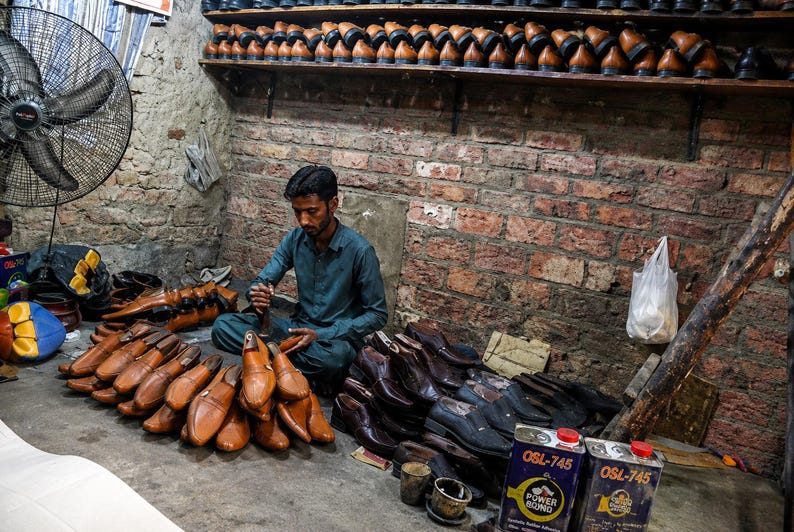 K&ouml;nnte beinhalten: Ein Schuhmacher sitzt auf dem Boden in einer Werkstatt, umgeben von zahlreichen hell- und dunkelbraunen Lederschuhen, Schuhleisten und zwei Metalldosen mit der Aufschrift OSL-745 Power Bond. Regale voller Lederschuhe s&auml;umen die Ziegelwand.