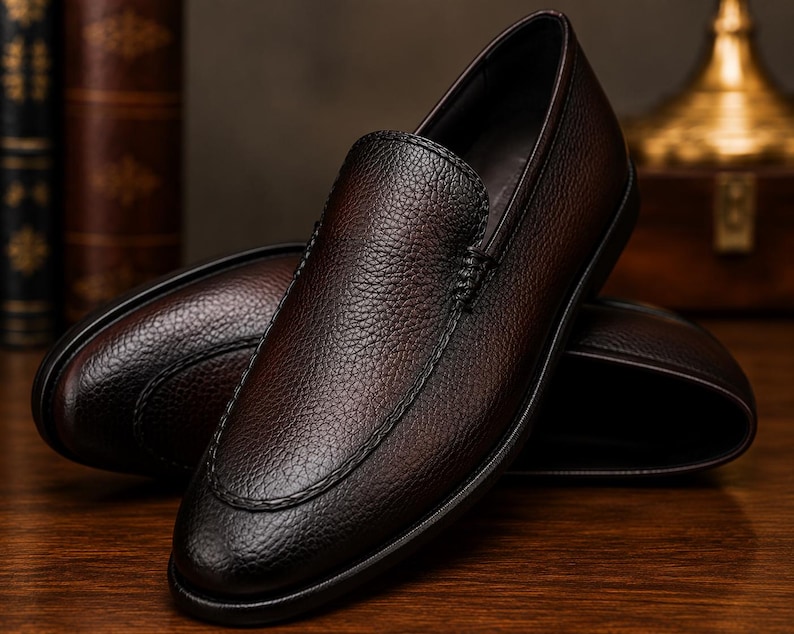 May include: A pair of dark brown loafers featuring a textured, pebbled leather upper with visible stitching along the vamp. The shoes have a rounded toe, a low-profile silhouette, and a smooth, dark sole, presenting a clean and simple design.