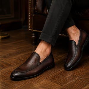 May include: A pair of dark brown leather loafers with a textured, pebbled finish and a rounded toe. The shoes feature a slip-on design with visible stitching along the vamp and a low, flat black sole, paired with dark trousers.