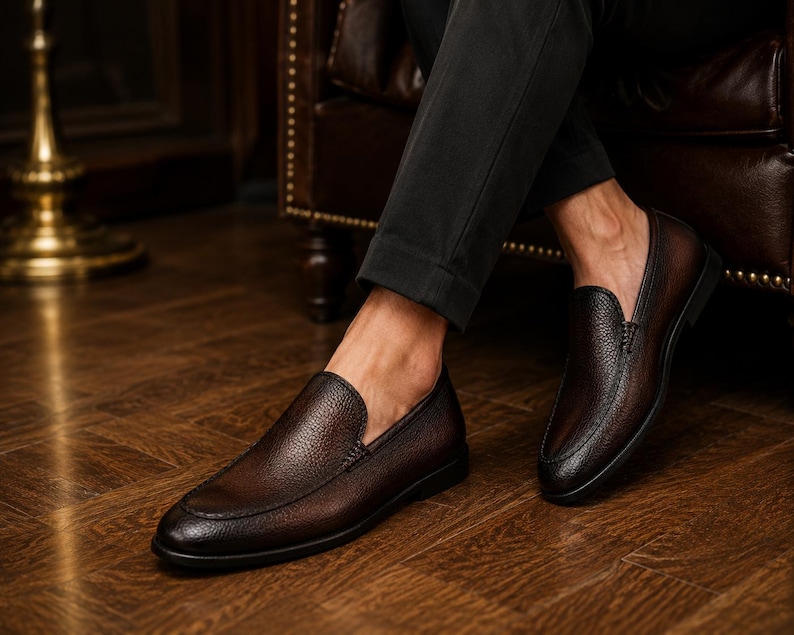 May include: A pair of dark brown leather loafers with a textured, pebbled finish and a rounded toe. The shoes feature a slip-on design with visible stitching along the vamp and a low, flat black sole, paired with dark trousers.