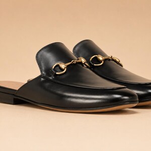May include: A pair of black leather backless loafers featuring a smooth finish, a rounded toe, and a gold-toned metal horsebit detail across the vamp. The shoes have a low heel and a tan-colored interior lining, showcasing a slip-on design.