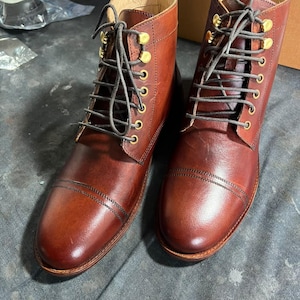 Puede incluir: Un par de botas de cuero marrón con puntera reforzada, con cierre de cordones gris oscuro y ojales metálicos dorados. Las botas tienen un acabado de cuero liso, una puntera redondeada con costuras dobles y un forro interior de color claro.