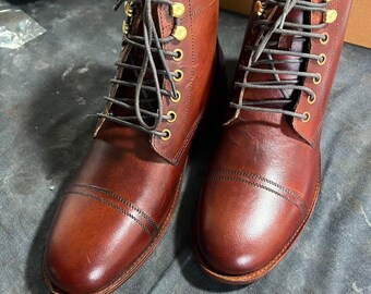 Könnte beinhalten: Ein Paar braune Lederstiefel mit Zehenkappe, ausgestattet mit einer Schnürung mit dunkelgrauen Schnürsenkeln und goldfarbenen Metallösen. Die Stiefel haben eine glatte Lederoberfläche, eine abgerundete Zehenkappe mit Doppelnaht und ein helles Innenfutter.