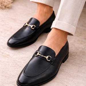 K&ouml;nnte beinhalten: Ein Paar schwarze Leder-Loafer mit einem goldfarbenen Metall-Trensendetail &uuml;ber dem Vorderblatt. Die Schuhe haben eine glatte Oberfl&auml;che, eine abgerundete Zehenpartie und einen niedrigen Absatz, kombiniert mit einer hellen Hose.