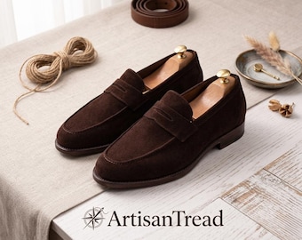 Könnte beinhalten: Ein Paar dunkelbraune Wildleder-Loafer mit Holzschuhspannern. Sie werden auf einer hellen Oberfläche präsentiert, zusammen mit einer Garnrolle, einem Ledergürtel und einer kleinen Schale mit dekorativen Elementen. Das ArtisanTread-Logo ist sichtbar.