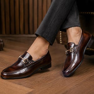 K&ouml;nnte beinhalten: Ein Paar dunkelbraune Leder-Loafer mit poliertem Finish, abgerundeter Schuhspitze und einem goldfarbenen Metall-Trensendetail &uuml;ber dem Vorderblatt. Die Schuhe haben einen niedrigen Absatz und eine glatte, strukturierte Form mit sichtbaren N&auml;hten.
