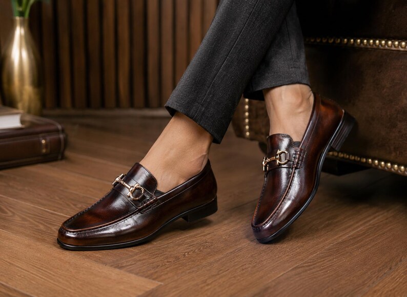 K&ouml;nnte beinhalten: Ein Paar dunkelbraune Leder-Loafer mit poliertem Finish, abgerundeter Schuhspitze und einem goldfarbenen Metall-Trensendetail &uuml;ber dem Vorderblatt. Die Schuhe haben einen niedrigen Absatz und eine glatte, strukturierte Form mit sichtbaren N&auml;hten.