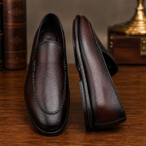 May include: A pair of dark brown loafers with a textured, pebbled leather finish. One shoe is positioned upright showing the top view, while the other is placed on its side to display the profile and the dark sole. The design features stitching.