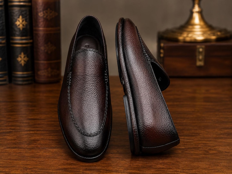 May include: A pair of dark brown loafers with a textured, pebbled leather finish. One shoe is positioned upright showing the top view, while the other is placed on its side to display the profile and the dark sole. The design features stitching.