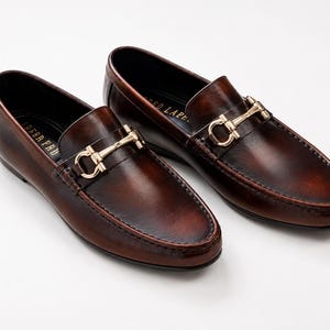 K&ouml;nnte beinhalten: Ein Paar dunkelbraune Leder-Loafer mit poliertem Finish, abgerundeter Zehenpartie und einem goldfarbenen Metall-Trensendetail &uuml;ber dem Vorderblatt. Die schwarze Innensohle zeigt den goldenen Schriftzug "IDEE PELLE" im Fersenbereich.