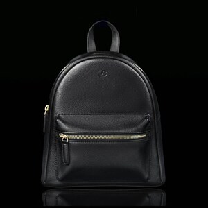 Puede incluir: Una mochila negra con textura granulada, que cuenta con un asa superior, un compartimento principal con cremallera y un bolsillo frontal más pequeño con cremallera. El bolsillo frontal tiene un tirador de metal dorado y un logo grabado arriba.