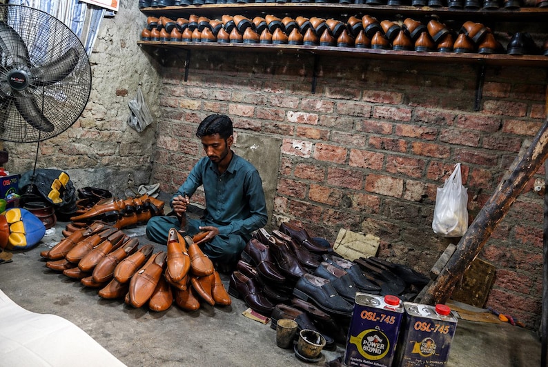 May include: A shoemaker sits on the floor in a workshop surrounded by numerous tan and dark brown leather shoes, shoe lasts, and two metal cans labeled OSL-745 Power Bond. Shelves filled with rows of finished leather footwear line the brick wall.