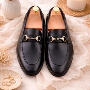 K&ouml;nnte beinhalten: Ein Paar schwarze Leder-Loafer mit einem goldfarbenen Metall-Trensendetail &uuml;ber dem Vorderblatt. Jeder Schuh enth&auml;lt einen hellen Holz-Schuhspanner, der im Inneren eingesetzt ist, um die Form des Schuhwerks zu erhalten. Die Loafer haben eine glatte Oberfl&auml;che.
