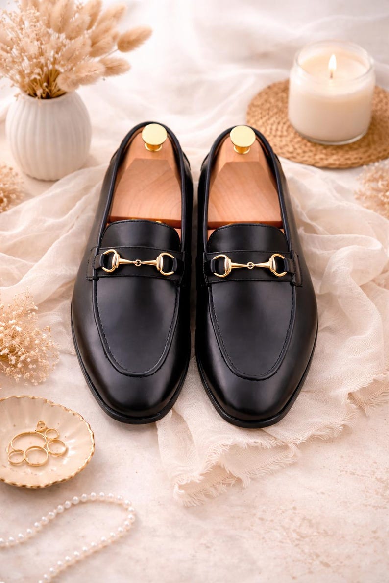 K&ouml;nnte beinhalten: Ein Paar schwarze Leder-Loafer mit einem goldfarbenen Metall-Trensendetail &uuml;ber dem Vorderblatt. Jeder Schuh enth&auml;lt einen hellen Holz-Schuhspanner, der im Inneren eingesetzt ist, um die Form des Schuhwerks zu erhalten. Die Loafer haben eine glatte Oberfl&auml;che.