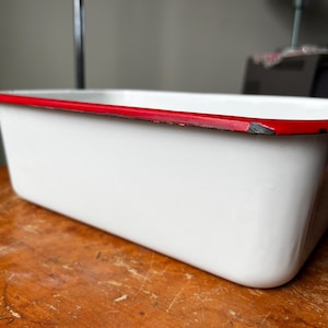 Puede incluir: Un recipiente rectangular de esmalte blanco con un borde rojo. Esta pieza de menaje de cocina vintage se encuentra sobre una superficie de madera. La pintura roja muestra signos de desgaste, lo que añade encanto antiguo.