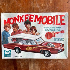 Puede incluir: Caja de modelo de coche Monkeemobile vintage. La caja presenta una ilustración de un coche granate con el texto "MONKEEMOBILE" en letras rojas grandes. La caja también incluye imágenes de los miembros de la banda Monkees y el texto "1/25 SCALE KIT".