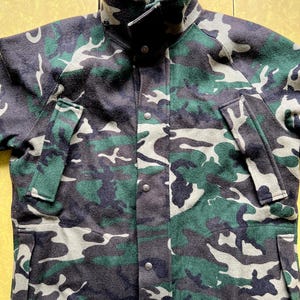 Puede incluir: Chaqueta de camuflaje en tonos verdes, grises y negros. La chaqueta tiene cuello alto, cierre de botones y múltiples bolsillos. La tela tiene un aspecto texturizado. El patrón es militar.