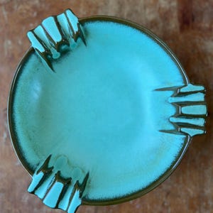 May include: A turquoise ceramic ashtray with a brown rim and three built-in rests. The ashtray has a round shape with a smooth, glossy interior. The rests are textured and have a dark brown colour.