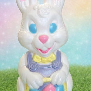 Könnte beinhalten: Eine weiße Osterhasenfigur mit rosa Innenohren, blauen Augen und einer rosa Nase und Mund. Der Hase trägt eine lila Latzhose und eine gelbe Fliege und hält einen gelben Korb mit blauen und rosa Ostereiern.