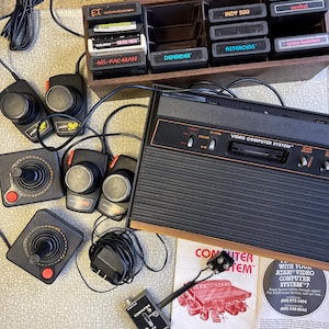 May include: An Atari Video Computer System with joysticks, paddle controllers, and game cartridges. Cartridges include "E.T.", "Ms. Pac-Man", "Defender", "Asteroids", and "Space Invaders". The system includes the owner's manual and a TV adapter.