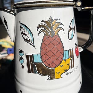 May include: White enamelware coffee pot with a black handle and lid. The pot is decorated with a large pineapple, leaves, and geometric shapes in gold, brown, blue, and red. The pot has a gold rim and the name "Georges Briard" is printed on the side.