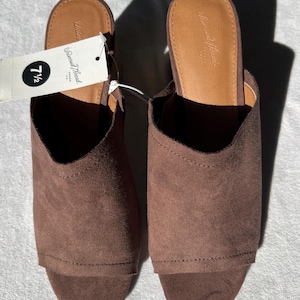 May include: A pair of brown suede slip-on mules. The shoes have a rounded toe and a low heel. A tag attached to the left shoe displays the size 7 1/2. The brand name "Universal Thread" is printed on the insole.