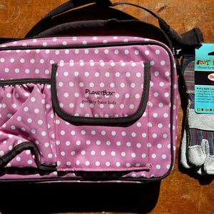 Pode incluir: Uma lancheira PlanetBox rosa com bolinhas brancas, alça e acabamentos pretos. Ao lado, um par de luvas de trabalho infantis com o texto "JUST FOR KIDS" na etiqueta.