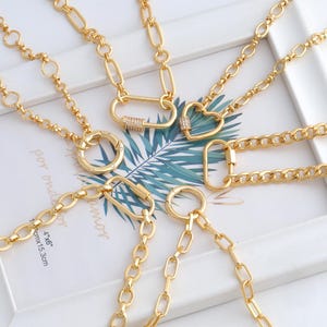 May include: A collection of gold-colored necklaces with various pendant designs, including a heart, a circle, and a carabiner clasp. The necklaces are displayed on a white surface with a tropical leaf print.