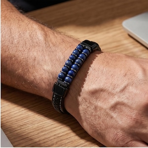 Puede incluir: Una pulsera de cuero negro con dos filas de cuentas azules y detalles plateados. La pulsera tiene un cierre negro y se lleva en la muñeca. Es un accesorio elegante.
