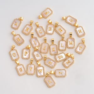 May include: Gold-colored initial pendants with a rectangular mother-of-pearl center and a border of small, clear crystals. Each pendant features a different letter of the alphabet, from A to Z, in a gold-colored font. The pendants are displayed on a light background.