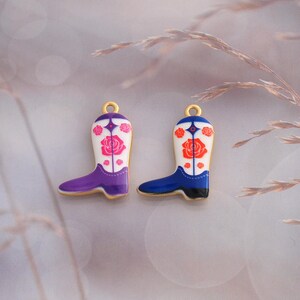 Puede incluir: Dos dijes de metal con forma de bota vaquera con cañas blancas y patrones de rosas. Una bota tiene detalles en morado y rosas rosadas, mientras que la otra tiene detalles en azul y rosas rojas. Cada dije incluye una pequeña argolla dorada arriba.