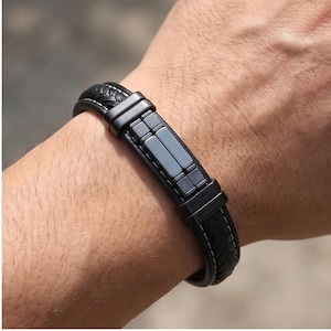 May include: Black leather bracelet with a braided design and a rectangular metal plate. The bracelet features white stitching and a secure clasp. A stylish accessory for everyday wear.