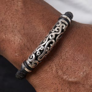 May include: A black leather bracelet with a silver, ornate, scrollwork design. The bracelet features a braided leather band and silver accents. The focal point is a cylindrical silver piece with intricate patterns. A stylish accessory.