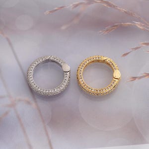 Puede incluir: Dos anillos circulares de resorte, uno en tono plateado y otro en tono dorado, ambos con una superficie pavé con numerosas piedras pequeñas, claras y brillantes incrustadas a lo largo de toda la circunferencia exterior y un mecanismo de bisagra.